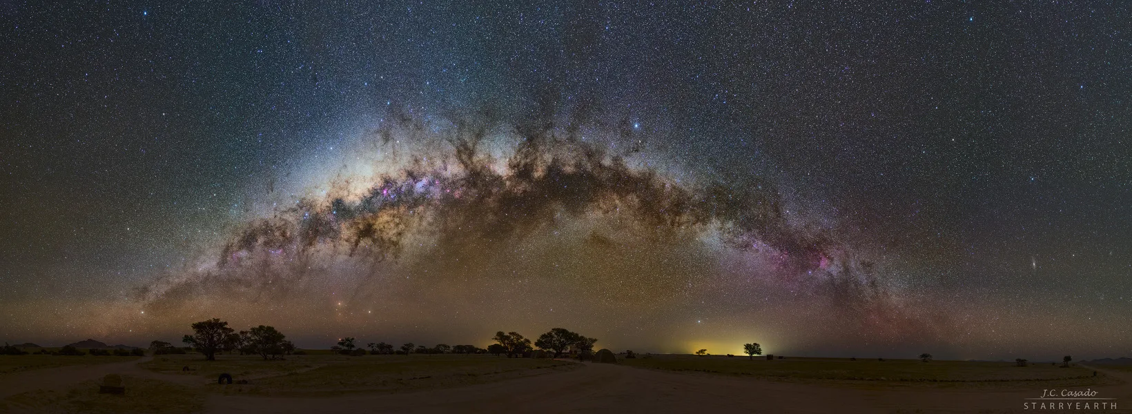 A 180° panorama of the Milky Way arching over the dark Namibian desert: a hazy band of countless stars, brightest and most bulging toward the Galactic centre, crossed by dark dust lanes, with the faint glow of distant towns on the horizon.